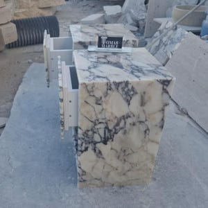 Puede incluir: Dos mesitas de noche de m&aacute;rmol con cajones blancos. Las mesas tienen un patr&oacute;n veteado blanco y gris. Los cajones est&aacute;n abiertos, revelando el interior. Un letrero que dice "TOMAS MARBLE" est&aacute; encima de una mesa.