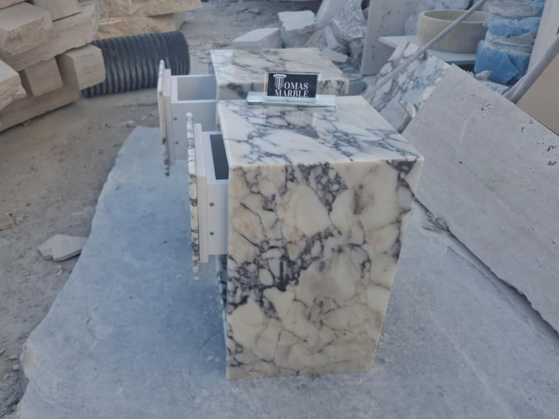 Puede incluir: Dos mesitas de noche de m&aacute;rmol con cajones blancos. Las mesas tienen un patr&oacute;n veteado blanco y gris. Los cajones est&aacute;n abiertos, revelando el interior. Un letrero que dice "TOMAS MARBLE" est&aacute; encima de una mesa.