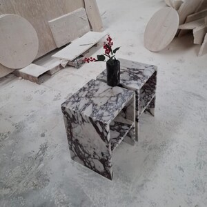 May include: Two matching side tables crafted from white marble with prominent dark purple veining. Each table features a rectangular top and a lower shelf. A black cylindrical vase containing a sprig of red berries sits on the top surface.