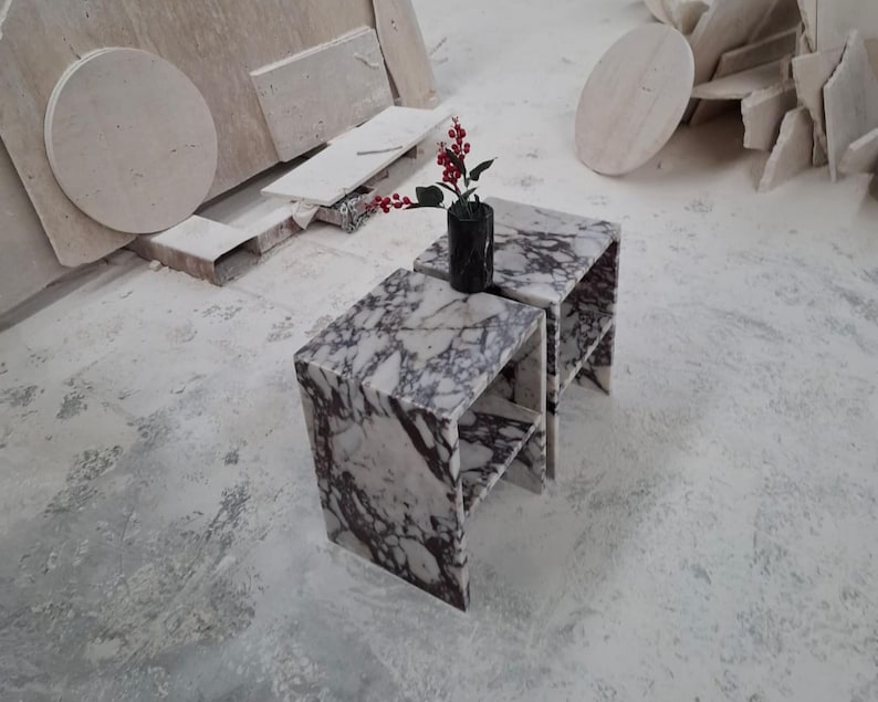 May include: Two matching side tables crafted from white marble with prominent dark purple veining. Each table features a rectangular top and a lower shelf. A black cylindrical vase containing a sprig of red berries sits on the top surface.