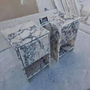 Puede incluir: Dos mesitas de noche de m&aacute;rmol con un patr&oacute;n veteado blanco y gris. Cada mesita de noche tiene un caj&oacute;n y una balda abierta. Una balda de cristal conecta las dos mesitas. El letrero de la balda de cristal dice "TOMAS MARBLE".