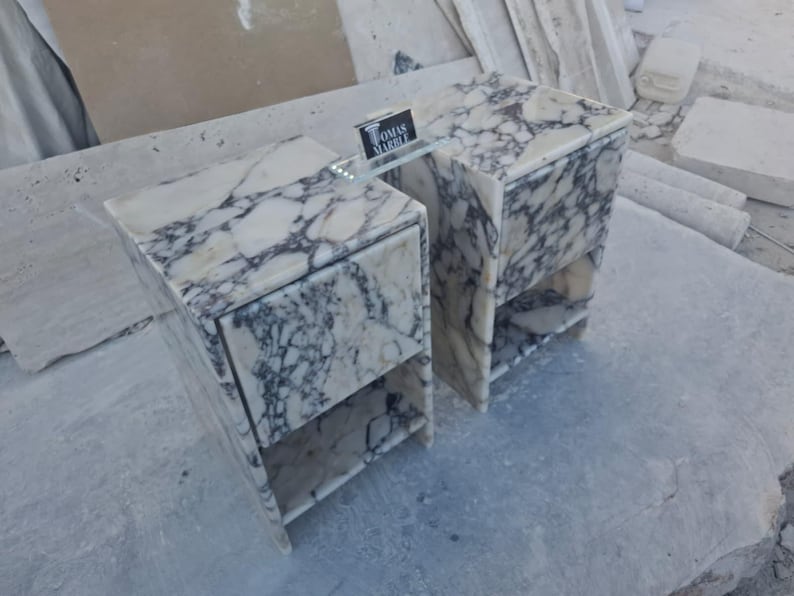 Puede incluir: Dos mesitas de noche de m&aacute;rmol con un patr&oacute;n veteado blanco y gris. Cada mesita de noche tiene un caj&oacute;n y una balda abierta. Una balda de cristal conecta las dos mesitas. El letrero de la balda de cristal dice "TOMAS MARBLE".