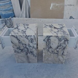Puede incluir: Dos mesitas de noche de m&aacute;rmol con cajones blancos. Las mesas son rectangulares con un patr&oacute;n veteado blanco y gris. Los cajones est&aacute;n extra&iacute;dos, revelando el interior blanco. Las mesas est&aacute;n sobre una superficie gris claro.