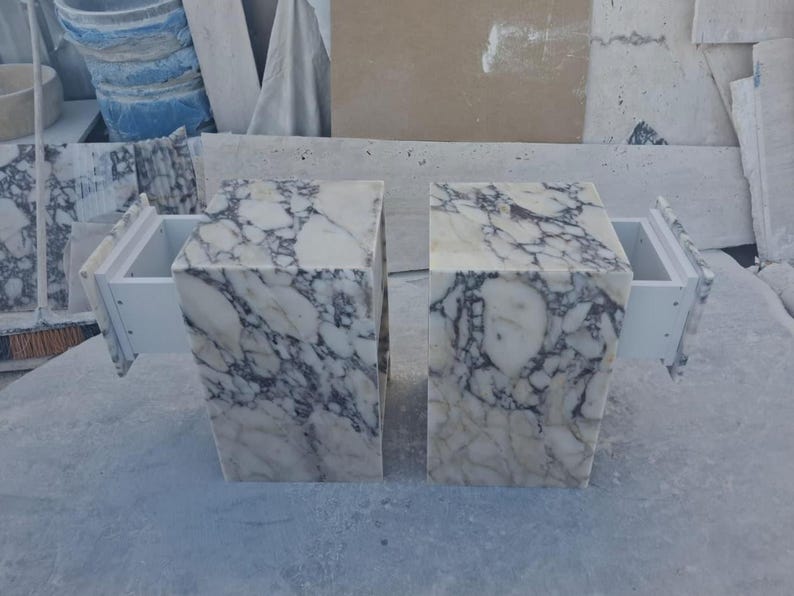 Puede incluir: Dos mesitas de noche de m&aacute;rmol con cajones blancos. Las mesas son rectangulares con un patr&oacute;n veteado blanco y gris. Los cajones est&aacute;n extra&iacute;dos, revelando el interior blanco. Las mesas est&aacute;n sobre una superficie gris claro.