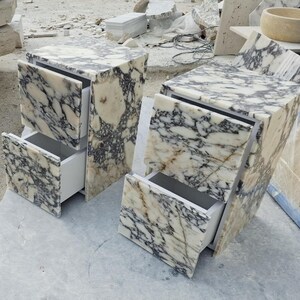 May include: Two bedside tables crafted from white and black veined marble. Each unit features a rectangular top and two drawers with matching marble fronts. The interior drawer boxes are white, and the marble displays a high-gloss polished finish.