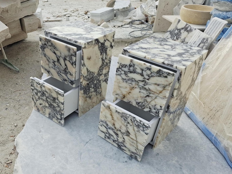 May include: Two bedside tables crafted from white and black veined marble. Each unit features a rectangular top and two drawers with matching marble fronts. The interior drawer boxes are white, and the marble displays a high-gloss polished finish.