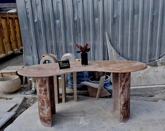 May include: A kidney-shaped console table crafted from brown marble with a polished surface and two thick, curved slab legs. A small black sign with the text TOMAS MARBLE sits on the tabletop next to a black vase containing red berries.