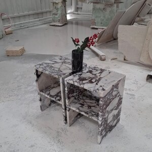 May include: Two matching side tables crafted from white marble with prominent dark purple veining. Each table features a rectangular top, a single open shelf, and a solid base. A black cylindrical vase containing red berries sits on top.