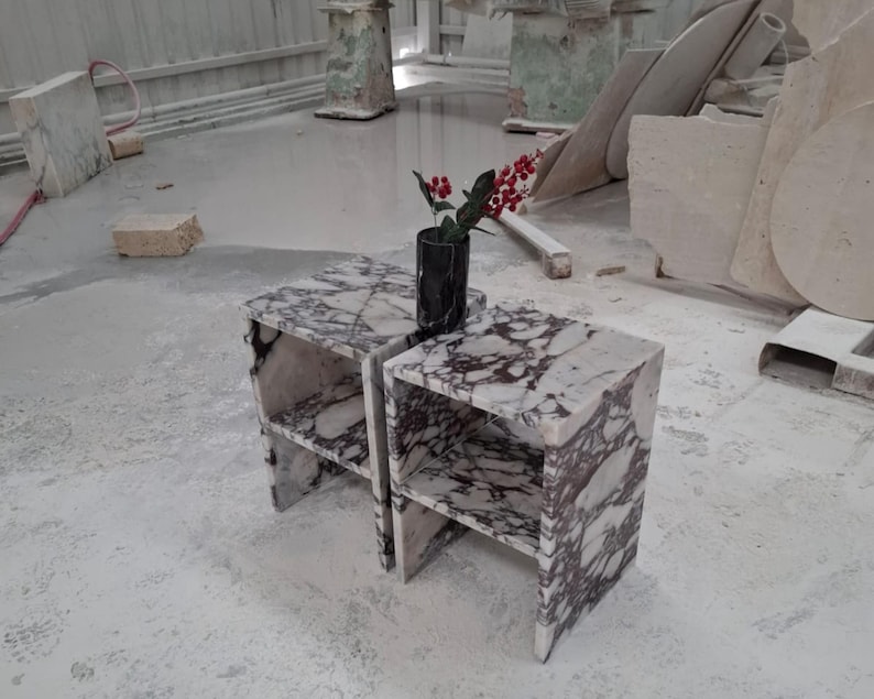 May include: Two matching side tables crafted from white marble with prominent dark purple veining. Each table features a rectangular top, a single open shelf, and a solid base. A black cylindrical vase containing red berries sits on top.
