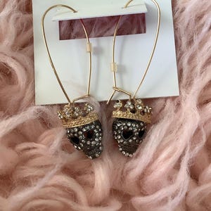 May include: Gold-tone drop earrings featuring skull pendants with rhinestone accents and small crowns. The earrings are attached to a white card with the brand name "Betsey Johnson" in red script.