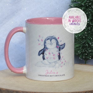 May include: A white ceramic mug with a pink rim and handle. The mug features a cartoon penguin holding pink and white Christmas lights. The text "Julia's Christmas Hot Chocolate" is printed on the mug.