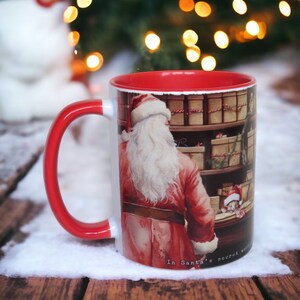 May include: A festive ceramic mug with a red interior and handle. The mug features an illustration of Santa Claus in a red suit, standing in front of shelves filled with wrapped gifts. The text "In Santa's secret world" is printed on the mug.