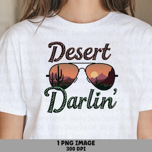 May include: A white t-shirt featuring a graphic of aviator sunglasses with a desert sunset scene inside the lenses. The word Desert is printed in dark red above the glasses, and the word Darlin' is printed in dark green below the glasses.