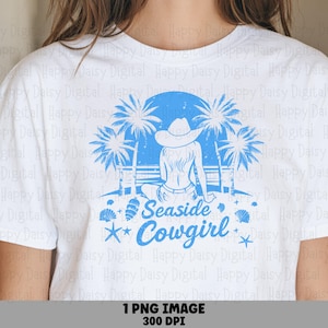 May include: A white t-shirt featuring a blue graphic of a person wearing a cowboy hat sitting on a beach with palm trees. The design includes the text "Seaside Cowgirl" in a script font, surrounded by small illustrations of seashells and starfish.