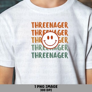 May include: A white short-sleeved t-shirt featuring the text THREENAGER printed in orange and green stacked rows. A large orange smiley face icon is centered over the middle row of text, partially obscuring the word THREENAGER.