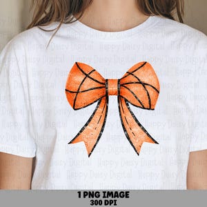 Può includere: Una t-shirt bianca a maniche corte con una grande grafica di un fiocco arancione con linee nere che ricordano la trama e il motivo di un pallone da basket. Il disegno è centrato sul petto del capo, che presenta un girocollo.