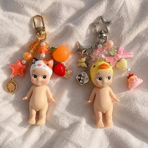 May include: Two keychains featuring small doll figures. The left keychain has a cat-eared hood, orange star, orange fruit, and strawberry charms on a gold clasp. The right keychain has a duck-hooded figure, star, dice, and cake charms on a silver star clasp.