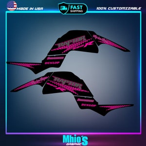 Puede incluir: Dise&ntilde;o gr&aacute;fico rosa y negro para un ATV Raptor 700R. El dise&ntilde;o presenta la palabra "RAPTOR" en una fuente estilizada con un patr&oacute;n de cuadros en blanco y negro. El dise&ntilde;o tambi&eacute;n incluye las palabras "DUNLOP" y "700R".