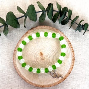 May include: A beaded bracelet featuring alternating translucent white and vibrant green faceted beads arranged in a circular pattern. A small silver-toned metal charm shaped like a chemical molecular structure hangs from the bracelet.