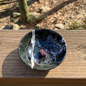 May include: A small, dark purple crystal sits in a black and white marble bowl. The bowl is on a wooden surface.