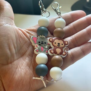 May include: Two beaded keychains featuring silicone beads and animal charms. One keychain has a gray elephant charm with pink ears and white and gray beads. The other has a brown monkey charm with brown and white beads. Both have silver metal clasps.