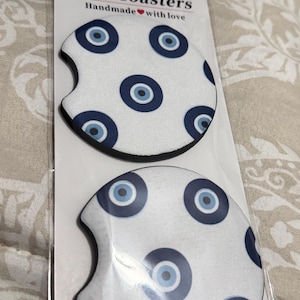 May include: Two car coasters with a white background and a pattern of blue evil eye designs. The coasters are packaged in a clear plastic sleeve. The text "Car Coasters Handmade with love" is printed on the packaging.
