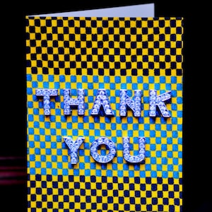 May include: A greeting card with a checkered pattern in yellow, blue, and black. The words "THANK YOU" are spelled out in silver, jewel-encrusted letters. The card is open, revealing a white interior.