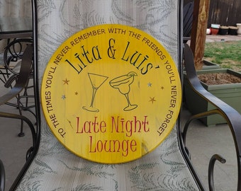 May include: A yellow wooden round sign with the text "You'll never remember with the friends you'll never forget" surrounding the text "Lita & Luis" and "Late Night Lounge". Two black and white illustrations of cocktail glasses are above the text "Late Night Lounge".
