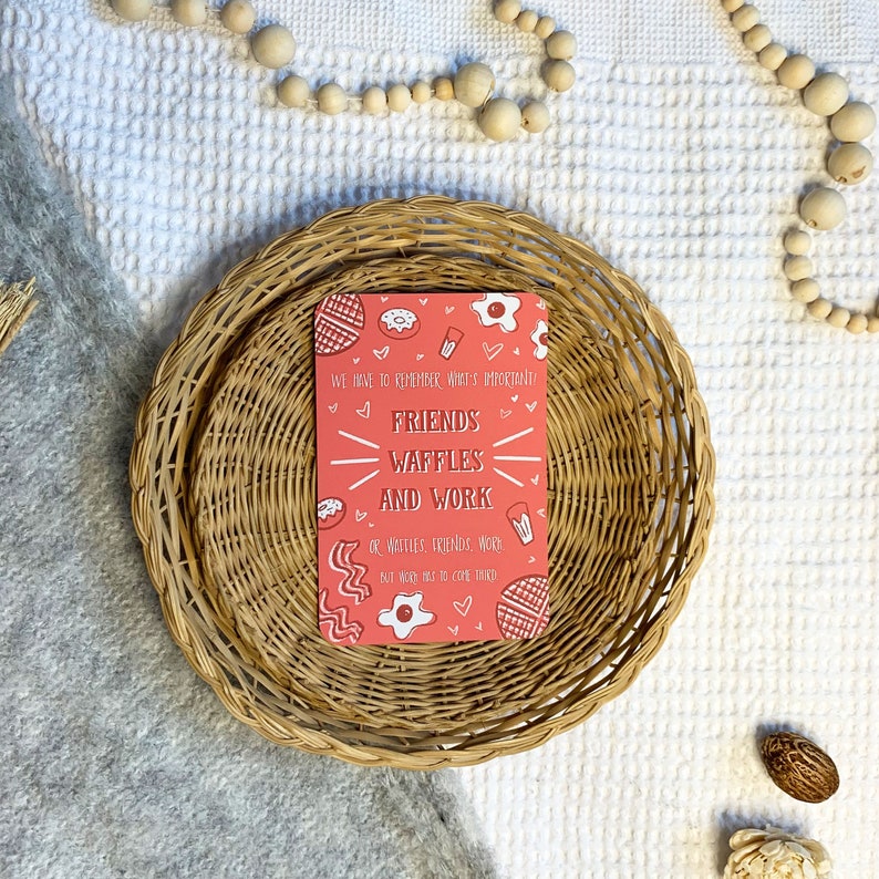 May include: A pink card with a white border and a message that reads "We have to remember what's important! Friends, Waffles and Work or Waffles, Friends, Work but no one has to core them". The card is decorated with hearts, waffles, eggs, bacon, and donuts.