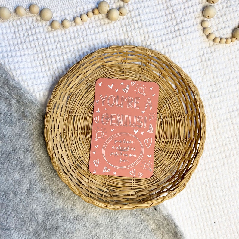 May include: A pink card with white text that reads "YOU'RE A GENIUS!" and "your brain is almost as perfect as your face." The card is surrounded by white hearts and light bulbs.