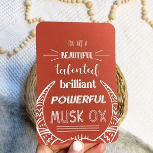 May include: A red card with white text that reads "You are a beautiful talented brilliant powerful musk ox". The card has a white illustration of a musk ox head with horns.