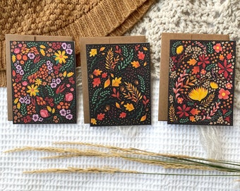 May include: Three cards with black backgrounds and colorful floral patterns. The cards are on a white surface with a brown knitted blanket in the background.