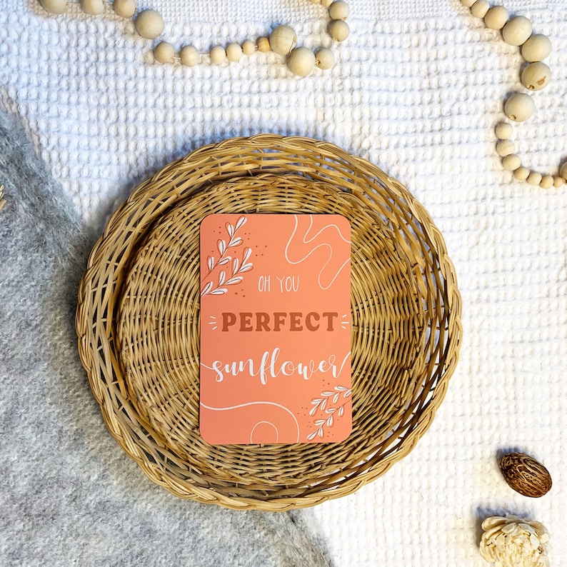 May include: A peach-colored card with white lettering and a floral design reads "Oh you perfect sunflower". The card is sitting on top of two woven wicker baskets.
