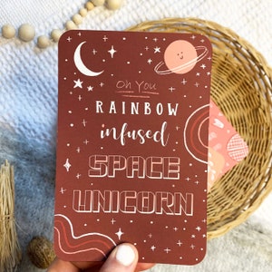 May include: A red card with white text that reads "Oh You Rainbow infused Space Unicorn". The card has a pink planet with a smiley face and a crescent moon with stars.