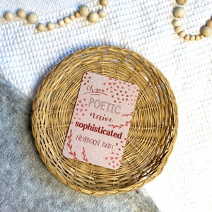 May include: A pink card with the text "Oh you POETIC naive sophisticated NEWBORN BABY" surrounded by small red and pink birds and leaves. The card is sitting in a woven wicker basket.