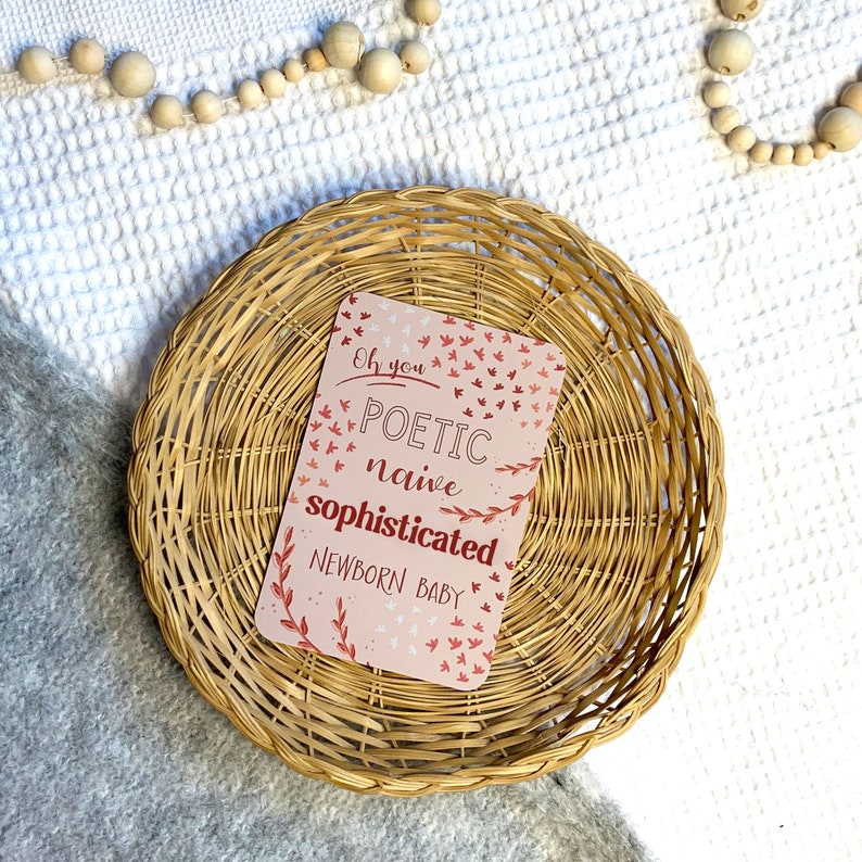 May include: A pink card with the text "Oh you POETIC naive sophisticated NEWBORN BABY" surrounded by small red and pink birds and leaves. The card is sitting in a woven wicker basket.