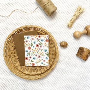 Puede incluir: Una tarjeta blanca con un patrón floral en tonos de rosa, azul, naranja y verde. La tarjeta está sentada sobre un sobre marrón en una cesta tejida.