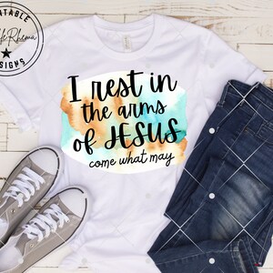 May include: White t-shirt with a watercolor design and the text "I rest in the arms of Jesus come what may".