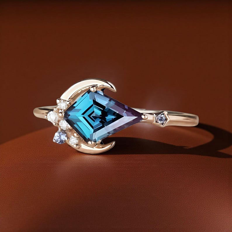 May include: A gold ring with a large, square-cut, teal gemstone with a purple hue. The ring has a crescent moon design on one side and small accent stones. A small star-shaped stone is set on the band.