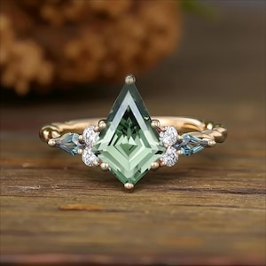 May include: A gold ring featuring a kite-shaped, green gemstone as the centerpiece. The ring is adorned with small, round white and blue gemstones. The band has a twisted rope design. The ring is set against a wooden surface.