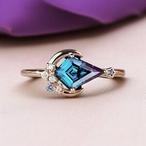 May include: A gold ring with a large, geometric, teal gemstone with a purple hue. The ring features a crescent moon design with small white and blue accent stones. A small star-shaped stone is set on the band.