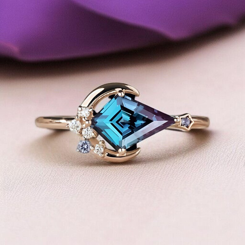 May include: A gold ring with a large, geometric, teal gemstone with a purple hue. The ring features a crescent moon design with small white and blue accent stones. A small star-shaped stone is set on the band.