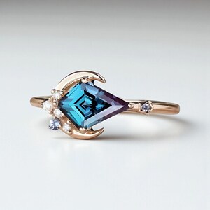 May include: A rose gold ring with a large, kite-shaped teal and purple gemstone. The ring features a crescent moon design and small accent stones. A small star-shaped stone is set on the band.