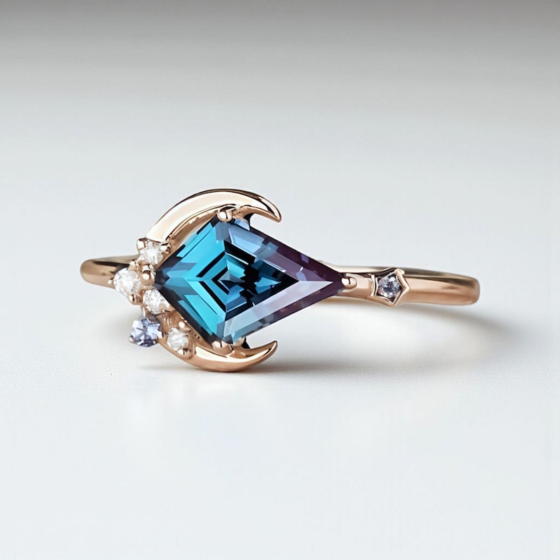 May include: A rose gold ring with a large, kite-shaped teal and purple gemstone. The ring features a crescent moon design and small accent stones. A small star-shaped stone is set on the band.