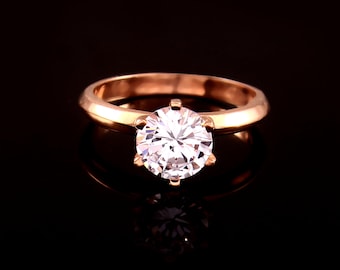May include: A rose gold engagement ring with a large, round, clear gemstone. The ring's band is a simple, polished design, and the gemstone is held in place by six prongs. The ring is set against a black background.