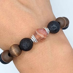 May include: A beaded bracelet featuring alternating dark brown wooden beads and black porous lava stone beads. The center of the bracelet showcases a single round, crackled orange stone bead flanked by two silver-toned metal spacer beads.