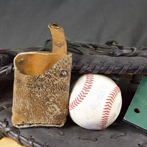 May include: A baseball-themed still life featuring a well-worn leather holster, a white baseball with red stitching, and a green number 5 sign, all resting on a black baseball glove. The holster is tan and has a textured pattern.