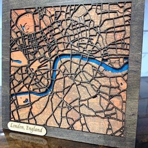 May include: A laser-cut wooden map of London, England, presented in a dark grey frame. The map showcases detailed streets and a blue river. The plaque at the bottom reads "London, England". A distinctive piece of wall art for the home.