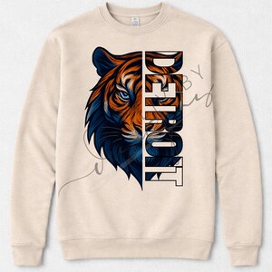 May include: A beige crew neck sweatshirt with a tiger graphic in orange and blue. The word "DETROIT" is printed vertically on the right side of the tiger's face. The design is centered on the front of the sweatshirt, which is a neutral colour.