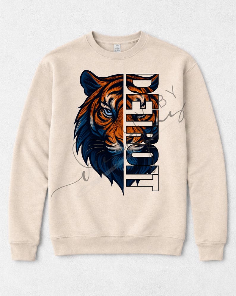 May include: A beige crew neck sweatshirt with a tiger graphic in orange and blue. The word "DETROIT" is printed vertically on the right side of the tiger's face. The design is centered on the front of the sweatshirt, which is a neutral colour.